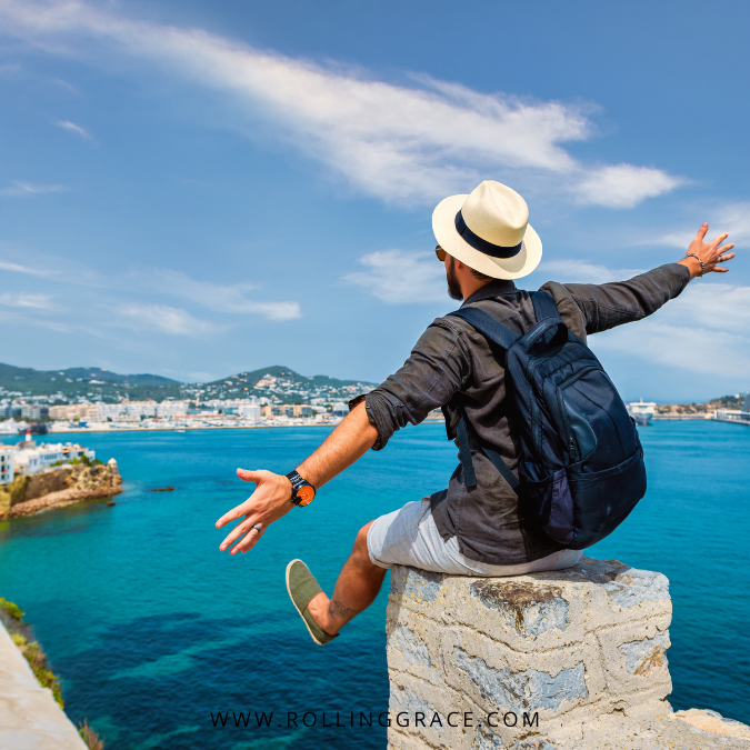 Traveller jumping with arms outstretched on a cliff overlooking a coastal town, overcoming travel anxiety