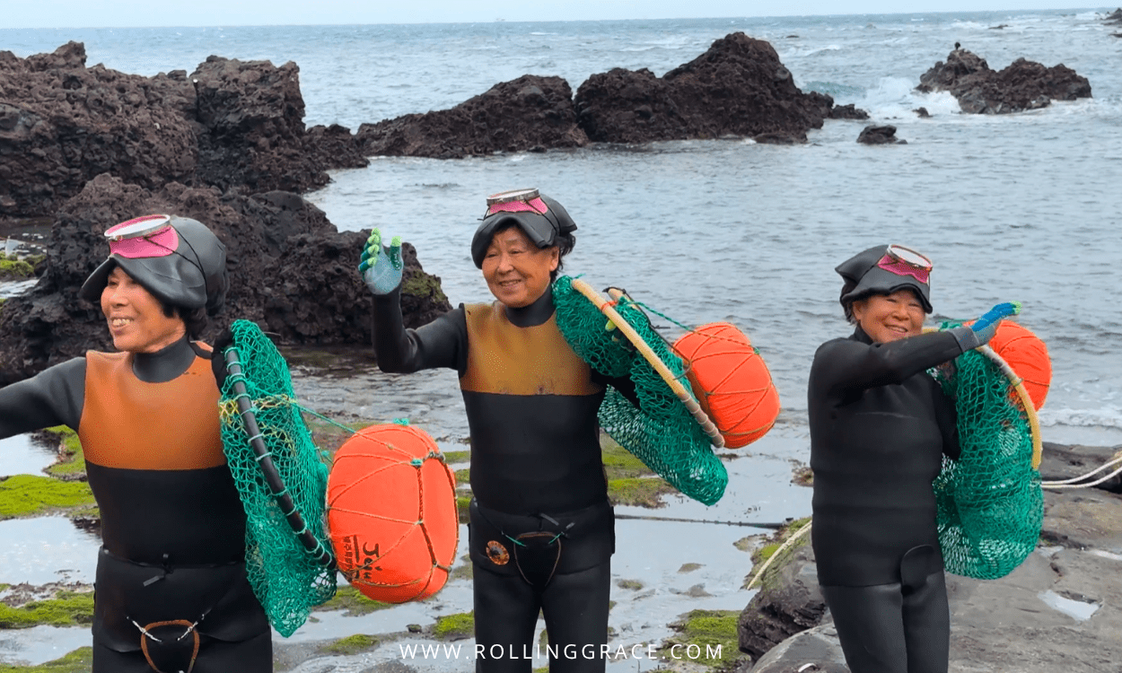 Three haenyeo divers on Jeju Island's volcanic rock coastline carrying orange floats and green nets after returning from a dive.