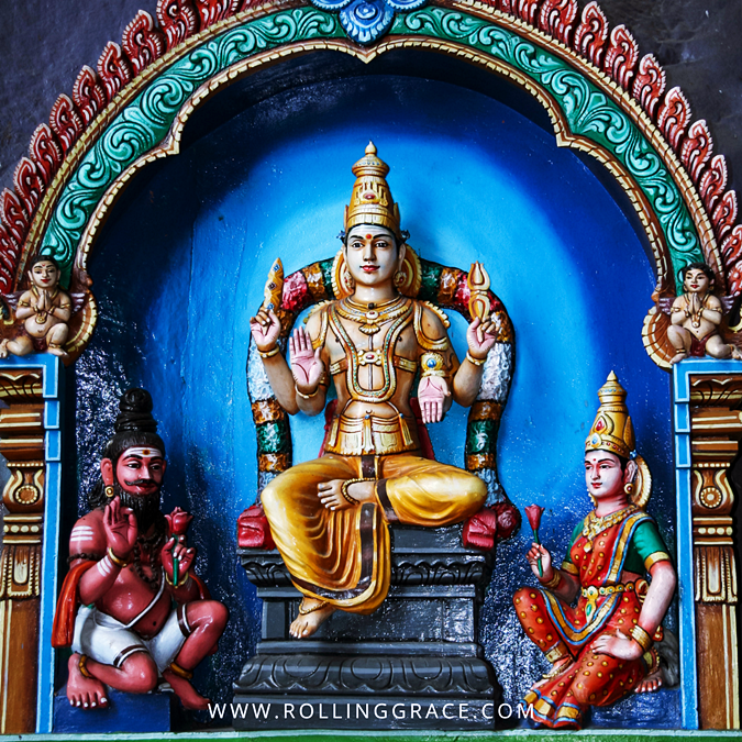 Painted shrine of a seated Hindu deity in a blue alcove, flanked by attendant figures, at Batu Caves