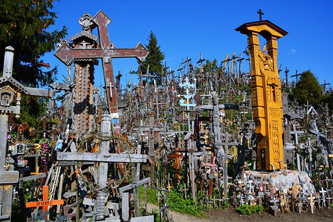 Hill of Crosses: 7 Must-See Sights and Incredible History 8 Tall carved orange wooden shrine pole and wooden crosses at the Hill of Crosses, Lithuania, against a blue sky
