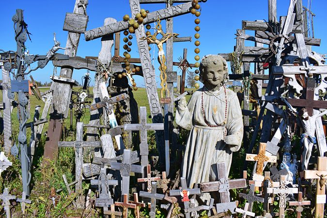 Hill of Crosses: 7 Must-See Sights and Incredible History 4 Carved statue of a robed figure among wooden and metal crosses at the Hill of Crosses, Lithuania, under a blue sky