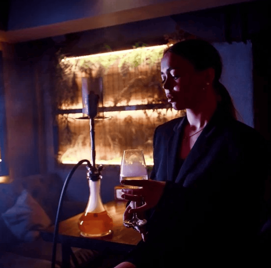 Guest holding a drink beside a hookah, lit by a glowing shelf with greenery at I'm Smoking, Arbatskaya, shisha in Moscow